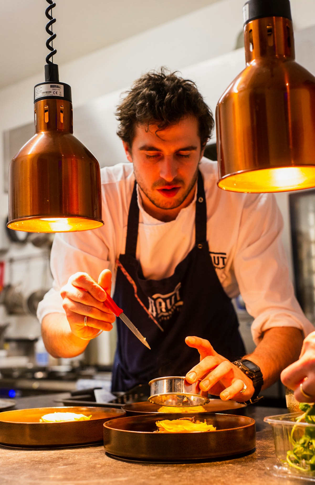 A chef plating a dish under copper heat lamps in a professional kitchen
